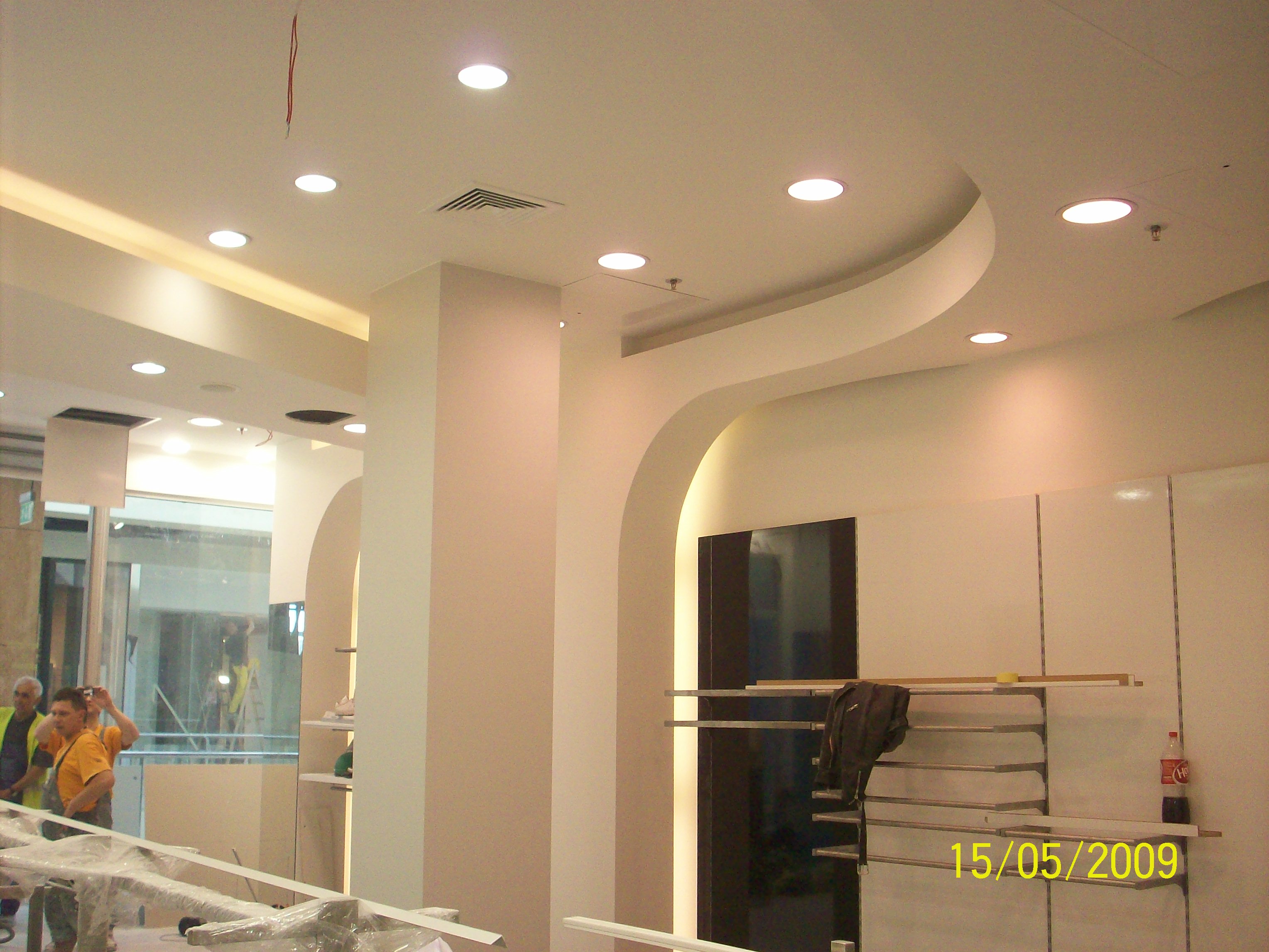 Wnętrze komercyjne w trakcie wykończenia: widoczne elementy zabudowy G/K, oświetlenie sufitowe, regały metalowe, pracownicy w tle, data na zdjęciu 15/05/2009.